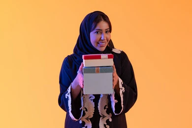 Interest in beauty, elegance, and appearance, holidays and happy occasions, celebration and gift-giving, a portrait of an Emirati Gulf Arab woman wearing a hijab and abaya holding a wrapped gift box in her hand, a Saudi woman standing with gestures of happiness and joy, the concept of femininity and softness, yellow background.