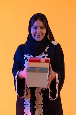 Holidays and happy occasions, celebration and gift-giving, a portrait of an Emirati Gulf Arab woman wearing a hijab and abaya holding a wrapped gift box in her hand, a Saudi woman standing with gestures of happiness and joy, the concept of femininity and softness, attention to beauty, elegance, and appearance, yellow background.