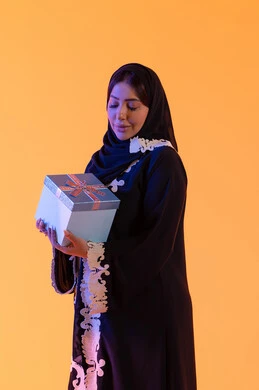 الأعياد والمناسبات السعيدة، الاحتفال وتقديم الهدايا، بورتريه لامرأة عربية خليجية اماراتيه ترتدي الحجاب والعباءة تحمل بيدها علبة هدية مغلفة ، سيده سعوديه تقف بايماءات السعادة والسرور، مفهوم الأنوثة والنعومة، الاهتمام بالجمال والأناقة والمظهر الخارجي، خلفية صفراء