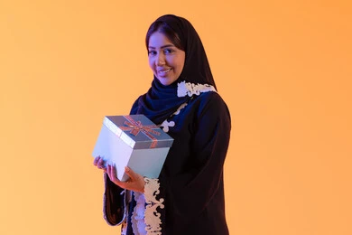 The concept of femininity and softness, holidays and happy occasions, celebration and gift-giving, a portrait of an Emirati Gulf Arab woman wearing a hijab and abaya holding a wrapped gift box in her hand, a Saudi woman standing with gestures of happiness and joy, attention to beauty, elegance, and appearance, yellow background.