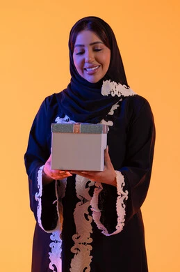بورتريه لامرأة عربية خليجية اماراتيه ترتدي الحجاب والعباءة تحمل بيدها علبة هدية مغلفة ، سيده سعوديه تقف بايماءات السعادة والسرور، مفهوم الأنوثة والنعومة، الاهتمام بالجمال والأناقة والمظهر الخارجي، الأعياد والمناسبات السعيدة، الاحتفال وتقديم الهدايا،خلفية صفراء