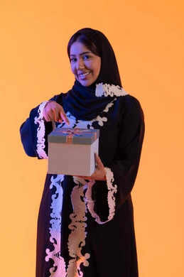 الاحتفال وتقديم الهدايا، الأعياد والمناسبات السعيدة، بورتريه لامرأة عربية خليجية اماراتيه ترتدي الحجاب والعباءة تحمل بيدها علبة هدية مغلفة ، سيده سعوديه تقف بايماءات السعادة والسرور، مفهوم الأنوثة والنعومة، الاهتمام بالجمال والأناقة والمظهر الخارجي، خلفية صفراء