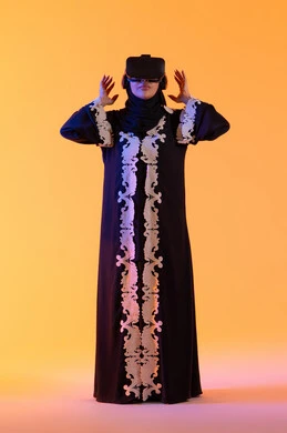 دمج التكنولوجيا بالحياة العملية، مفهوم الخوف والرعب ، محاولة الامساك بشئ ما، استخدام التقنيات الحديثة الالكترونية، شابة عربية خليجية اماراتية ترتدي العباءة المطرزة والحجاب تضع نظارات الفي آر ثلاثية الابعاد، سيدة سعوديه تقف بايماءات الاندماج والتفاعل مع العالم الافتراضي، خلفية صفراء