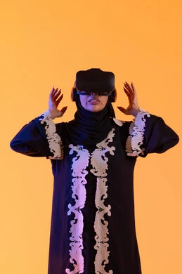 Attempting to grasp something, the concept of fear and terror, using modern electronic technologies, a young Arab Gulf Emirati woman wearing an embroidered abaya and hijab, putting on 3D VR glasses, a Saudi woman standing with gestures of integration and interaction with the virtual world, merging technology with practical life, yellow background.