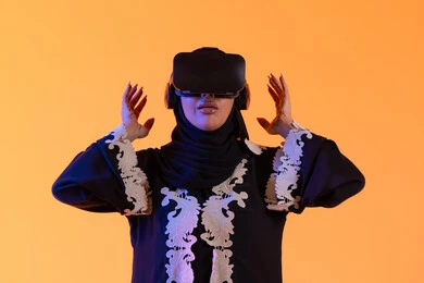 مفهوم الخوف والرعب ، محاولة الامساك بشئ ما، استخدام التقنيات الحديثة الالكترونية، شابة عربية خليجية اماراتية ترتدي العباءة المطرزة والحجاب تضع نظارات الفي آر ثلاثية الابعاد، سيدة سعوديه تقف بايماءات الاندماج والتفاعل مع العالم الافتراضي، دمج التكنولوجيا بالحياة العملية، خلفية صفراء