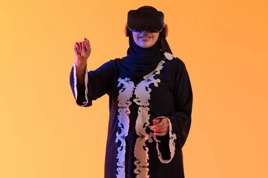 Integrating technology into practical life, attempting to click on something, using modern electronic technologies, a young Arab Gulf Emirati woman wearing an embroidered abaya and hijab, wearing 3D VR glasses, a Saudi woman standing with gestures of integration and interaction with the virtual world, yellow background.