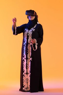 A young Arab Gulf Emirati woman wearing an embroidered abaya and hijab is wearing 3D VR glasses. A Saudi woman stands with gestures of integration and interaction with the virtual world, merging technology with practical life, trying to click on something, using modern electronic technologies, with a yellow background.
