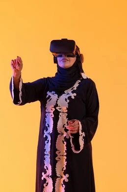 محاولة النقر علي شئ ما، استخدام التقنيات الحديثة الالكترونية، شابة عربية خليجية اماراتية ترتدي العباءة المطرزة والحجاب تضع نظارات الفي آر ثلاثية الابعاد، سيدة سعوديه تقف بايماءات الاندماج والتفاعل مع العالم الافتراضي، دمج التكنولوجيا بالحياة العملية، خلفية صفراء