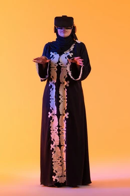 استخدام التقنيات الحديثة الالكترونية، مفهوم الخوف والرعب ، محاولة الامساك بشئ ما،شابة عربية خليجية اماراتية ترتدي العباءة المطرزة والحجاب تضع نظارات الفي آر ثلاثية الابعاد، سيدة سعوديه تقف بايماءات الاندماج والتفاعل مع العالم الافتراضي، دمج التكنولوجيا بالحياة العملية، خلفية صفراء