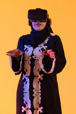 دمج التكنولوجيا بالحياة العملية، مفهوم الخوف والرعب ، محاولة الامساك بشئ ما، استخدام التقنيات الحديثة الالكترونية، شابة عربية خليجية اماراتية ترتدي العباءة المطرزة والحجاب تضع نظارات الفي آر ثلاثية الابعاد، سيدة سعوديه تقف بايماءات الاندماج والتفاعل مع العالم الافتراضي، خلفية صفراء