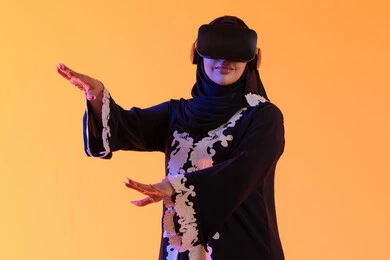 The use of modern electronic technologies, the concept of fear and horror, the attempt to grasp something, a young Arab Gulf Emirati woman wearing an embroidered abaya and hijab, wearing 3D VR glasses, a Saudi woman standing with gestures of integration and interaction with the virtual world, merging technology with practical life, yellow background.
