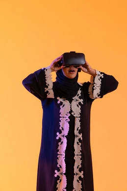 الاستمتاع في قضاء الأوقات الترفيهية عبر استخدام التقنيات الحديثة الالكترونية، شابة عربية خليجية سعودية محجبة ترتدي العباءة المطرزة والحجاب تضع نظارات الفي آر ثلاثية الابعاد، سيدة سعوديه تقف بايماءات الاندماج والتفاعل مع العالم الافتراضي، دمج التكنولوجيا بالحياة العملية، خلفية صفراء