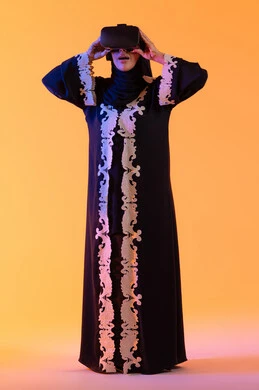 شابة عربية خليجية سعودية محجبة ترتدي العباءة المطرزة والحجاب تضع نظارات الفي آر ثلاثية الابعاد، سيدة سعوديه تقف بايماءات الاندماج والتفاعل مع العالم الافتراضي، دمج التكنولوجيا بالحياة العملية، الاستمتاع في قضاء الأوقات الترفيهية عبر استخدام التقنيات الحديثة الالكترونية، خلفية صفراء