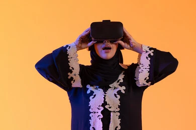 سيدة سعوديه تقف بايماءات الاندماج والتفاعل مع العالم الافتراضي، دمج التكنولوجيا بالحياة العملية، الاستمتاع في قضاء الأوقات الترفيهية عبر استخدام التقنيات الحديثة الالكترونية، شابة عربية خليجية اماراتية ترتدي العباءة المطرزة والحجاب تضع نظارات الفي آر ثلاثية الابعاد، خلفية صفراء