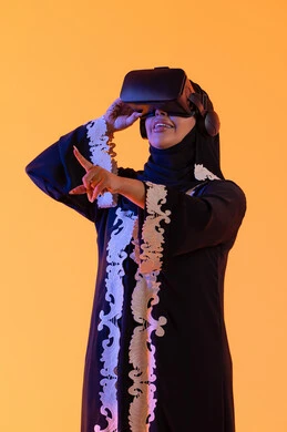 دمج التكنولوجيا بالحياة العملية، الاستمتاع في قضاء الأوقات الترفيهية عبر استخدام التقنيات الحديثة الالكترونية، شابة عربية خليجية سعودية محجبة ترتدي العباءة المطرزة والحجاب تضع نظارات الفي آر ثلاثية الابعاد، سيدة اماراتيه  تقف بايماءات الاندماج والتفاعل مع العالم الافتراضي، خلفية صفراء