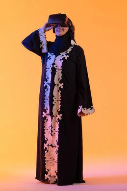 A young Arab Gulf Saudi woman wearing a decorated abaya and hijab, putting on 3D VR glasses, a Saudi lady standing with gestures of integration and interaction with the virtual world, merging technology with practical life, enjoying leisure time through the use of modern electronic technologies, with a yellow background.