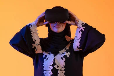 الاستمتاع في قضاء الأوقات الترفيهية عبر استخدام التقنيات الحديثة الالكترونية، شابة عربية خليجية اماراتيه ترتدي العباءة المطرزة والحجاب تضع نظارات الفي آر ثلاثية الابعاد، سيدة سعوديه تقف بايماءات الاندماج والتفاعل مع العالم الافتراضي، دمج التكنولوجيا بالحياة العملية، خلفية صفراء