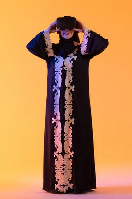 الاستمتاع في قضاء الأوقات الترفيهية عبر استخدام التقنيات الحديثة الالكترونية، شابة عربية خليجية اماراتية ترتدي العباءة المطرزة والحجاب تضع نظارات الفي آر ثلاثية الابعاد، سيدة سعوديه تقف بايماءات الاندماج والتفاعل مع العالم الافتراضي، دمج التكنولوجيا بالحياة العملية، خلفية صفراء