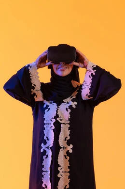 قضاء الأوقات الترفيهية عبر استخدام التقنيات الحديثة الالكترونية، شابة عربية خليجية سعودية محجبة ترتدي العباءة المطرزة والحجاب تضع نظارات الفي آر ثلاثية الابعاد، سيدة اماراتيه  تقف بايماءات الاندماج والتفاعل مع العالم الافتراضي، دمج التكنولوجيا بالحياة العملية