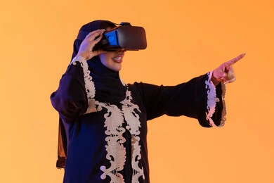 دمج التكنولوجيا بالحياة العملية، الاستمتاع في قضاء الأوقات الترفيهية عبر استخدام التقنيات الحديثة الالكترونية، شابة عربية خليجية سعودية محجبة ترتدي العباءة المطرزة والحجاب تضع نظارات الفي آر ثلاثية الابعاد، سيدة اماراتيه  تقف بايماءات الاندماج والتفاعل مع العالم الافتراضي