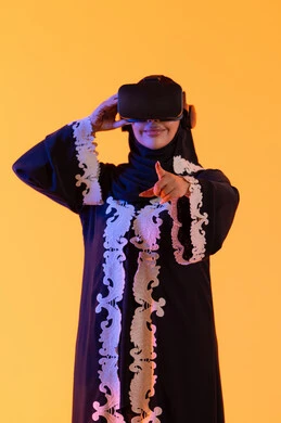 A young Arab Gulf Saudi woman wearing a decorated abaya and hijab, putting on 3D VR glasses, stands with gestures of integration and interaction with the virtual world, merging technology with practical life, enjoying leisure time through the use of modern electronic technologies.