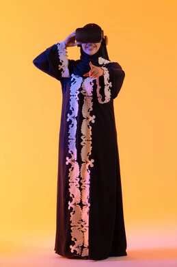 الاستمتاع في قضاء الأوقات الترفيهية عبر استخدام التقنيات الحديثة الالكترونية، شابة عربية خليجية سعودية محجبة ترتدي العباءة المطرزة والحجاب تضع نظارات الفي آر ثلاثية الابعاد، سيدة سعوديه تقف بايماءات الاندماج والتفاعل مع العالم الافتراضي، دمج التكنولوجيا بالحياة العملية