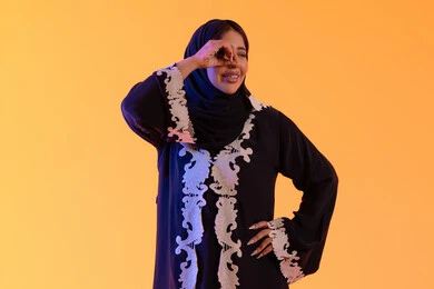 Looking with expressions of joy and enjoyment, seeing distant things and bringing them closer, a Saudi woman who loves exploration and adventure, a portrait of an Arab Gulf Emirati woman wearing an abaya and hijab, raising her hand with gestures of binoculars.