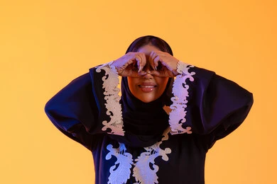 بورتريه لامراه عربيه خليجيه اماراتيه ترتدي العباءه والحجاب ترفع  يدها بايماءات منظار وتنظر بتعابير السرور والاستمتاع، رؤية الاشياء البعيدة وتقريبها، سيده سعوديه تحب الاستكشاف والمغامرة، خلفية صفراء