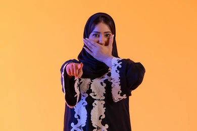 Expressions indicate this place here, specifying a certain location, a Saudi woman covering her mouth with her hand in a surprised expression, a portrait of an Emirati Gulf Arab woman wearing an abaya and hijab pointing with her index finger upwards to something, looking at the camera with gestures of joy and happiness.
