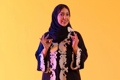 التعبير عن الجودة والاتقان، بورتريه لامراه عربيه خليجيه اماراتيه ترتدي العباءه والحجاب تقوم برفع ايديها بإيماءات الجودة والامتياز والتميز،سيده سعوديه تشير بتعابير الرضي والموافقة ، البشاشة والشعور بالسعادة ،  خلفية صفراء