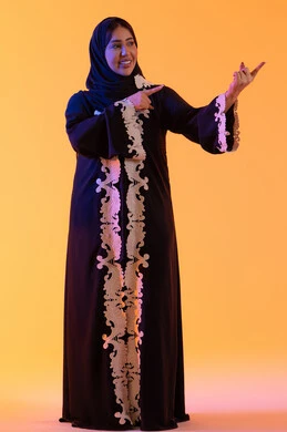 A portrait of an Arab Gulf Emirati woman wearing an abaya and hijab, pointing with her index finger upwards to something, looking at the camera with gestures of joy and happiness, expressions indicating this place here, specifying a certain location, with a yellow background.