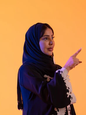 تعابير تشير هذا المكان هنا، تحديد موقع معين ، بورتريه لامراه عربيه خليجيه اماراتيه ترتدي العباءه والحجاب تشير بإصبع السبابة لاعلى إلى شيء ، النظر إلى الكاميرا بإيماءات الفرح والسرور، خلفية صفراء