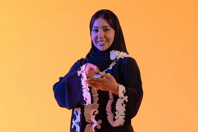 A portrait of an Arab Gulf Emirati woman wearing an embroidered abaya and hijab, raising her hand to display a holographic element and looking at the camera, with gestures of joy and happiness, three-dimensional hologram technology, a Saudi woman indicating by holding and containing a holographic element, with a yellow background.
