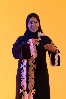 A Saudi woman is pointing while holding and containing a hologram element, a portrait of an Arab Gulf Emirati woman wearing an embroidered abaya and hijab, raising her hand to display a holographic element and looking at the camera, gestures of joy and happiness, three-dimensional hologram technology, yellow background.