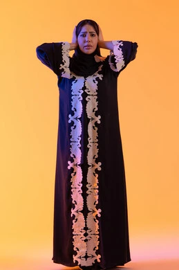 Astonishment and disbelief, loss and deprivation of something specific, surprise and wonder at something, a Saudi woman standing with gestures of amazement and astonishment, a portrait of an Arab Gulf Emirati woman wearing an embroidered abaya and hijab, raising her hands to her head with expressions of shock and bewilderment, yellow background.