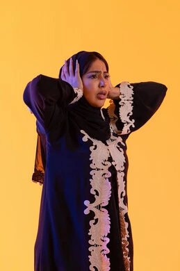 خسارة وفقدان شيء محدد, الانبهار وعدم التصديق,التفاجؤ والاستغراب من شيء ما, سيده سعوديه تقف بإيماءات التعجب والدهشة، بورتريه لامراه عربيه خليجيه اماراتيه ترتدي العباءه المطرزه والحجاب ترفع يديها على رأسه بتعابير الصدمة والذهول، خلفية صفراء