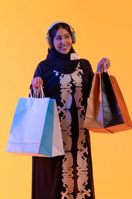 Shopping and purchasing from global brands, listening to music through headphones, the positive impact of shopping on women, a Saudi woman benefiting from seasonal offers and discounts, a portrait of a smiling Emirati Gulf Arab woman wearing an abaya and hijab holding colorful shopping bags in her hand, with a yellow background.