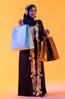 Using headphones, shopping and purchasing from global brands, the positive impact of shopping on women, a Saudi woman benefiting from seasonal offers and discounts, a portrait of a smiling Emirati Gulf Arab woman wearing an abaya and hijab holding colorful shopping bags in her hand, with a yellow background.