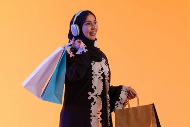 بورترية  لسيده عربية خليجية اماراتيه مبتسمة ترتدي العباءه والحجاب تحمل بيدها أكياس التسوق الملونه، سماع الموسيقي عبر الهيدفون ، التبضع والتسوق من الماركات العالميه، الأثر الإيجابي للتسوق على السيدات، سيدة سعودية تستفيد من العروض الموسمية والتخفيضات،خلفية صفراء