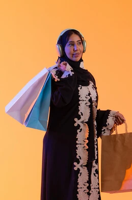 سماع الموسيقي عبر الهيدفون ، التبضع والتسوق من الماركات العالميه، الأثر الإيجابي للتسوق على السيدات، سيدة سعودية تستفيد من العروض الموسمية والتخفيضات، بورترية  لسيده عربية خليجية اماراتيه مبتسمة ترتدي العباءه والحجاب تحمل بيدها أكياس التسوق الملونه، ، خلفية صفراء