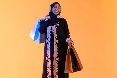 التبضع والتسوق من الماركات العالميه، بورترية لسيده عربية خليجية اماراتيه مبتسمة ترتدي العباءه والحجاب تحمل بيدها أكياس التسوق الملونه، الأثر الإيجابي للتسوق على السيدات، سيدة سعودية تستفيد من العروض الموسمية والتخفيضات،  خلفية صفراء