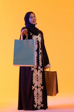سيدة سعودية تستفيد من العروض الموسمية والتخفيضات،  التبضع والتسوق من الماركات العالميه، بورترية لسيده عربية خليجية اماراتيه مبتسمة ترتدي العباءه والحجاب تحمل بيدها أكياس التسوق الملونه، الأثر الإيجابي للتسوق على السيدات،  خلفية صفراء