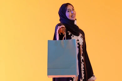 التبضع والتسوق من الماركات العالميه، بورترية لسيده عربية خليجية اماراتيه مبتسمة ترتدي العباءه والحجاب تحمل بيدها أكياس التسوق الملونه، الأثر الإيجابي للتسوق على السيدات، سيدة سعودية تستفيد من العروض الموسمية والتخفيضات،  خلفية صفراء