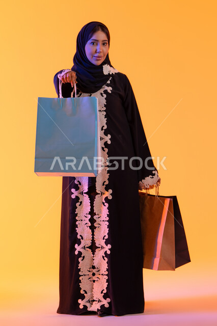 White Friday discounts, the positive impact of shopping on women, a Saudi woman benefiting from seasonal offers and discounts, shopping from global brands, a portrait of a smiling Emirati Gulf Arab woman wearing an abaya and hijab holding colorful shopping bags in her hand, yellow background.