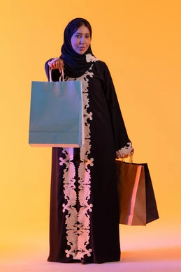 White Friday discounts, the positive impact of shopping on women, a Saudi woman benefiting from seasonal offers and discounts, shopping from global brands, a portrait of a smiling Emirati Gulf Arab woman wearing an abaya and hijab holding colorful shopping bags in her hand, yellow background.