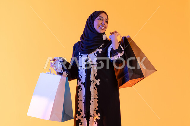 The positive impact of shopping on women, a Saudi woman benefiting from seasonal offers and discounts, shopping from global brands, a portrait of a smiling Arab Gulf Emirati woman wearing an abaya and hijab holding colorful shopping bags in her hand, with a yellow background.