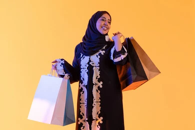 The positive impact of shopping on women, a Saudi woman benefiting from seasonal offers and discounts, shopping from global brands, a portrait of a smiling Arab Gulf Emirati woman wearing an abaya and hijab holding colorful shopping bags in her hand, with a yellow background.