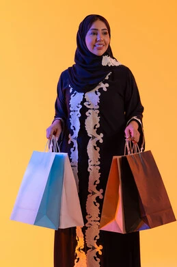 بورترية لسيده عربية خليجية اماراتيه مبتسمة ترتدي العباءه والحجاب تحمل بيدها أكياس التسوق الملونه، الأثر الإيجابي للتسوق على السيدات، سيدة سعودية تستفيد من العروض الموسمية والتخفيضات،  التبضع والتسوق من الماركات العالميه، خلفية صفراء