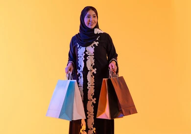 خصومات الجمعة البيضاء ، الأثر الإيجابي للتسوق على السيدات، سيدة سعودية تستفيد من العروض الموسمية والتخفيضات،  التبضع والتسوق من الماركات العالميه، بورترية لسيده عربية خليجية اماراتيه مبتسمة ترتدي العباءه والحجاب تحمل بيدها أكياس التسوق الملونه، ، خلفية صفراء