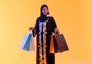 تعابير السرور والسعادة، الأثر الإيجابي للتسوق على السيدات، سيدة سعودية تستفيد من العروض الموسمية والتخفيضات،  التبضع والتسوق من الماركات العالميه، بورترية لسيده عربية خليجية اماراتيه مبتسمة ترتدي العباءه والحجاب تحمل بيدها أكياس التسوق الملونه، ، خلفية صفراء